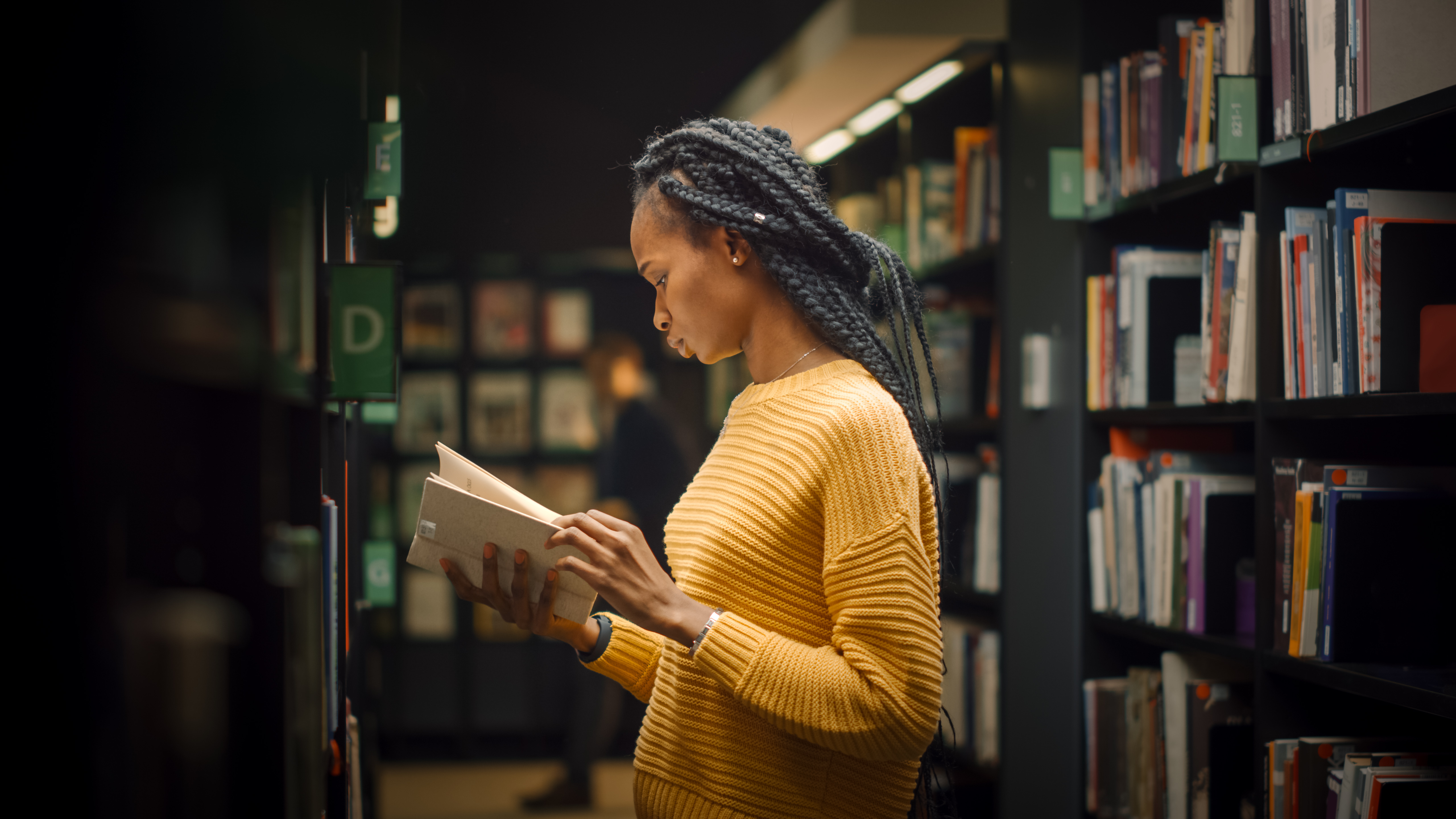 University Library: Portrait of Gifted Beautiful Black Girl Stands Between Rows of Bookshelves and Searching for the Right Book Title, Finds and Picks one for Class Assignment