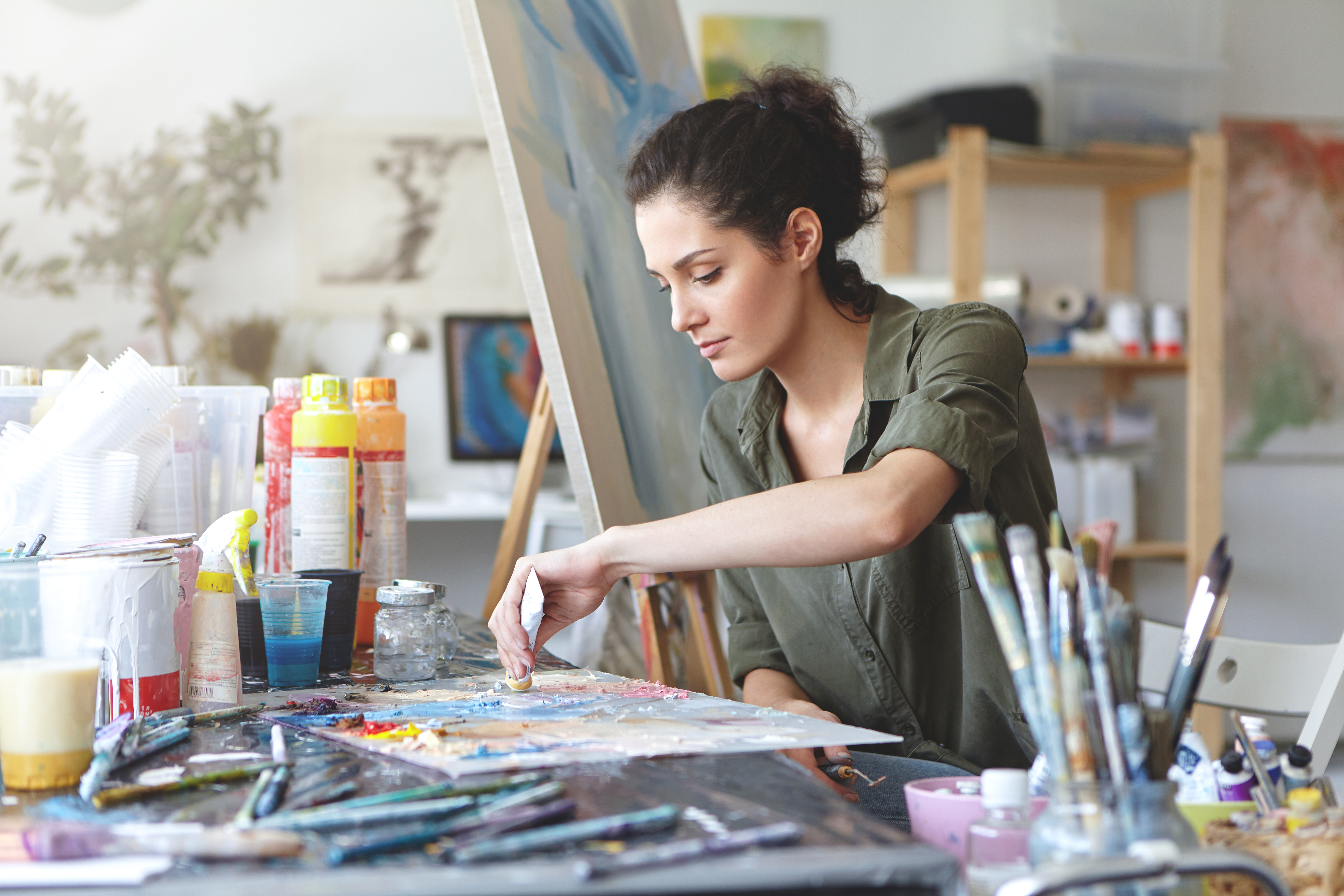 Serious brunette young beautiful woman sitting in art studio, taking colorful paints from tube while creating great masterpiece on easel, being preoccupied with her work, having nice imagination