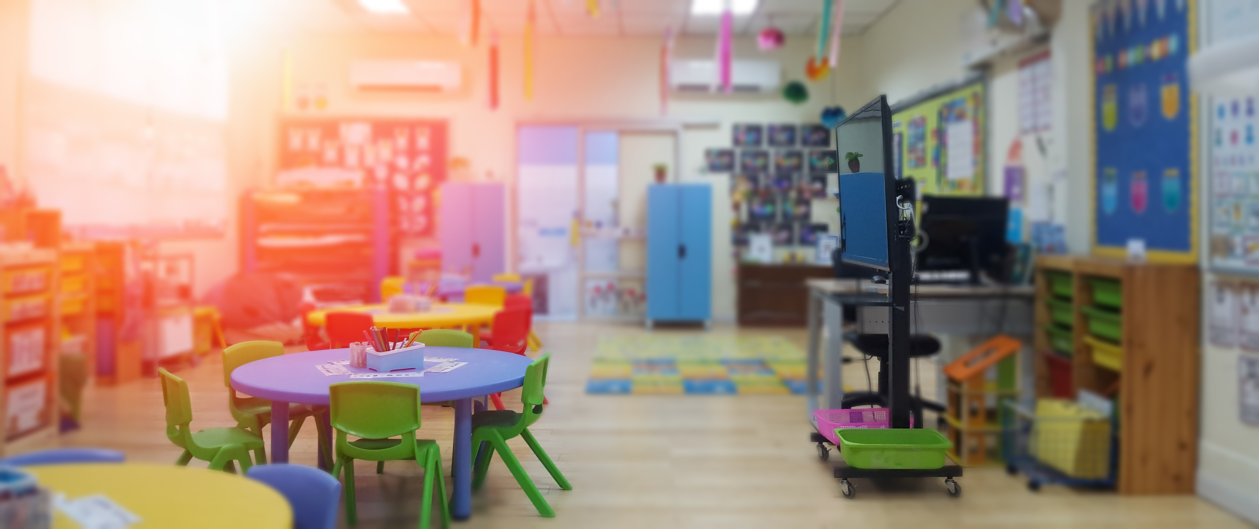 Early childhood classroom with small tables and chairs, play mats and teaching monitor.