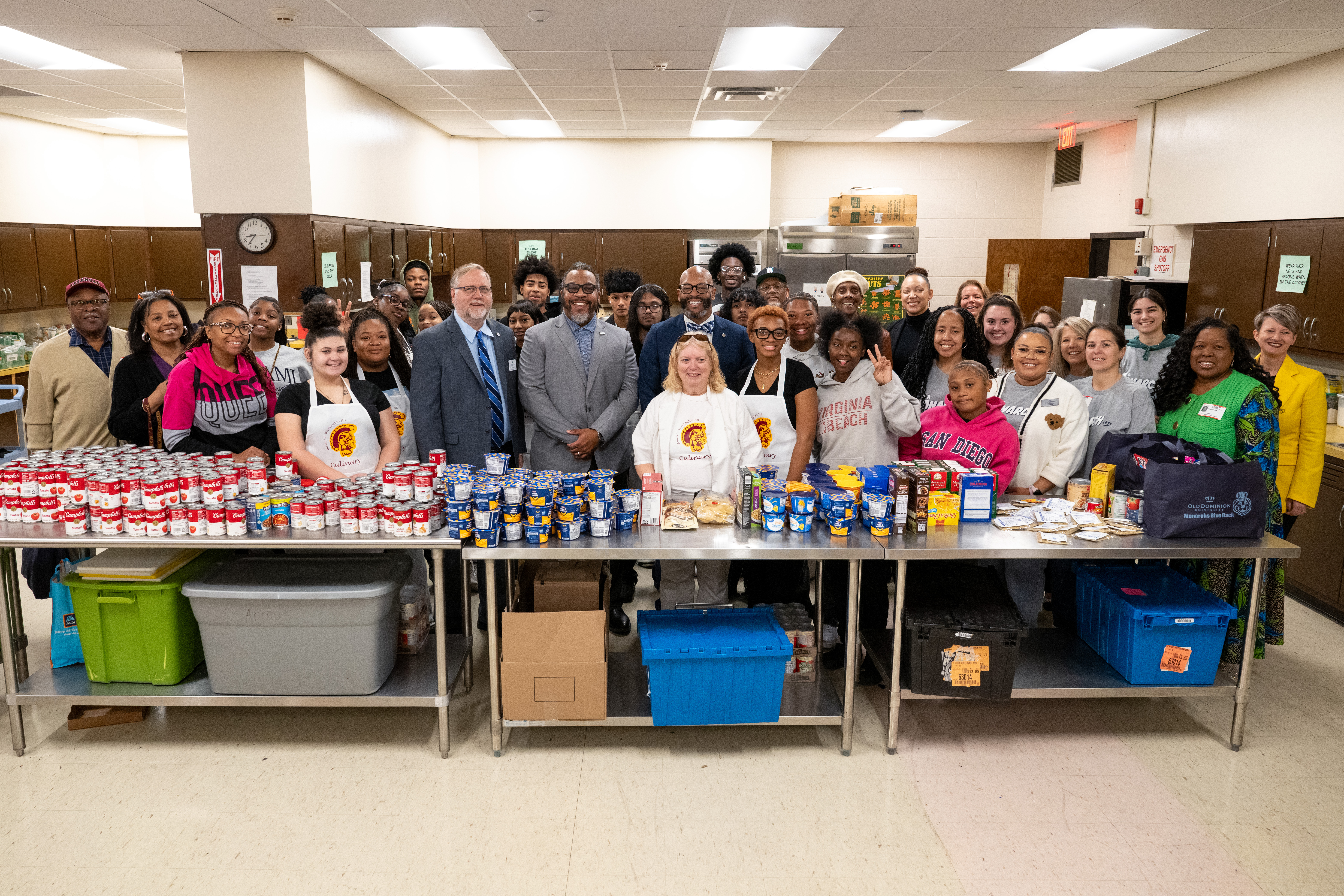 Officials and students from ODU and Norfolk Public Schools stand behind table in cafeteria kitchen with donated canned goods on top.