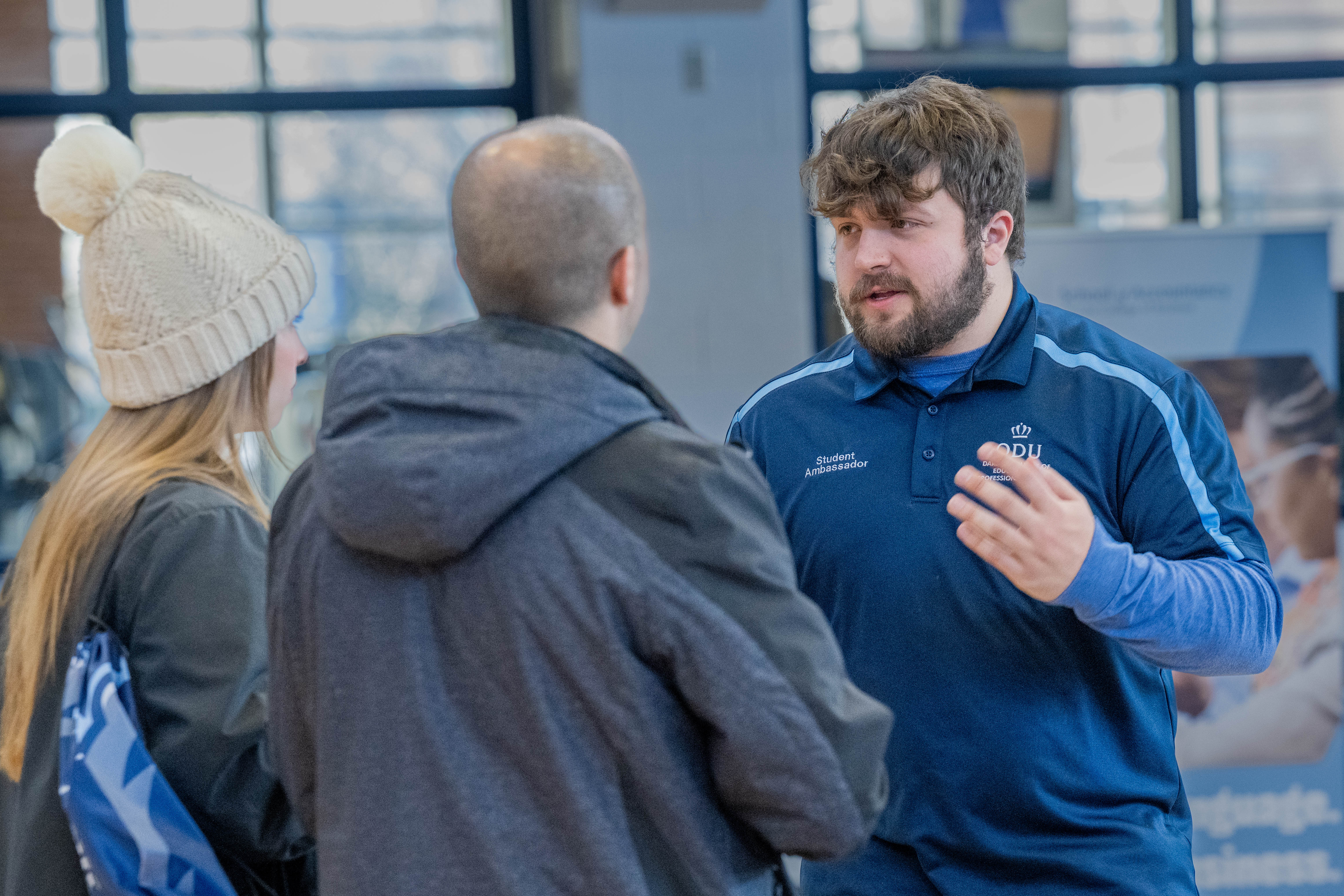 Adyn, wearing a blue collared shirt reading "Student Ambassador" talks to two visitors to campus, who are facing away from the camera.