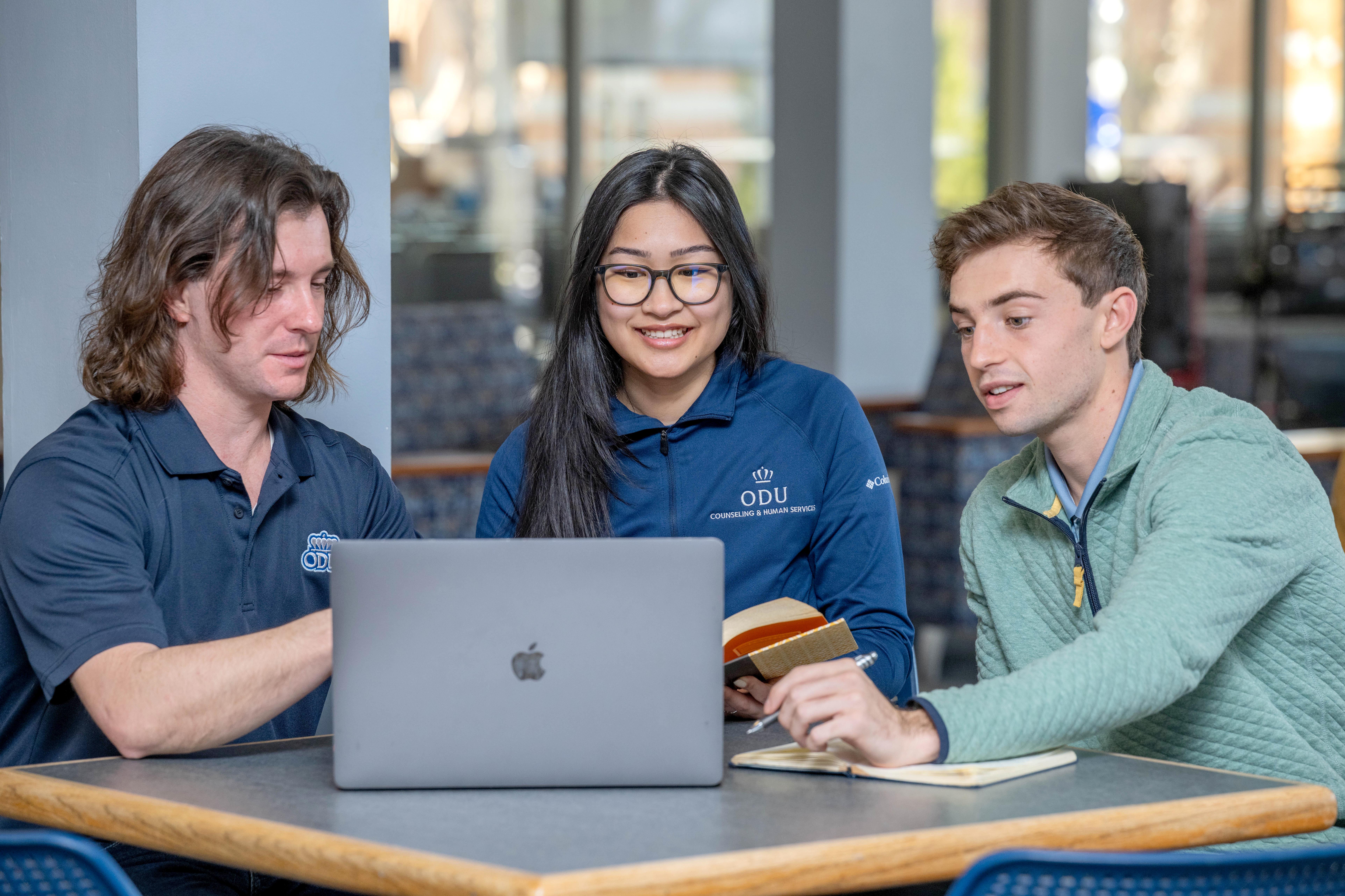 Three students sit at table looking at laptop.