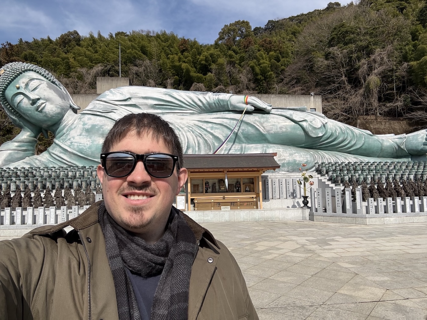 Dr. Melusky, wearing sunglasses, is pictured standing in front of a large reclining jade-colored figure while visiting the Nanzoin Temple in Sasaguri in the Fukuoka Prefecture. 