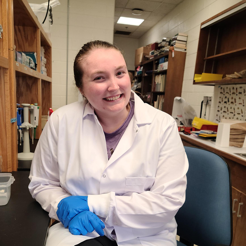 Emily Carey in a lab coat and gloves working in a lab.