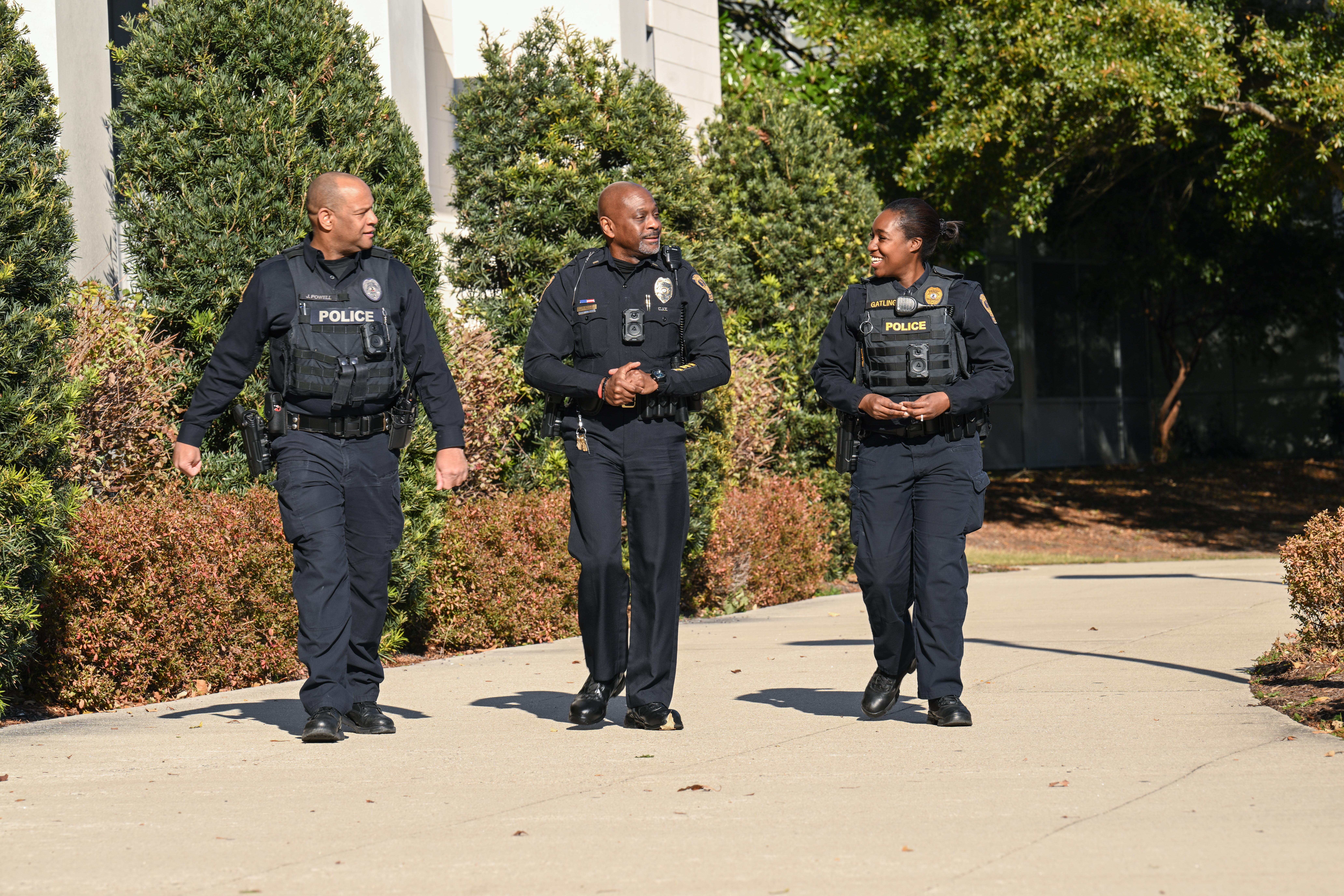 Three police officers in uniform walk down sidewalk while smiling.