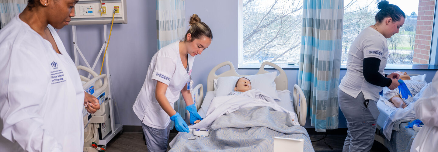 Three nursing students wearing white scrubs tend to mannequin patients in hospital simulation room.
