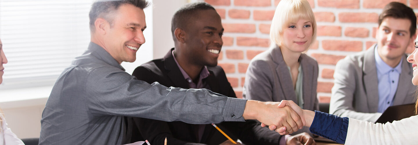 Female shakes hand of male as others the table smile.
