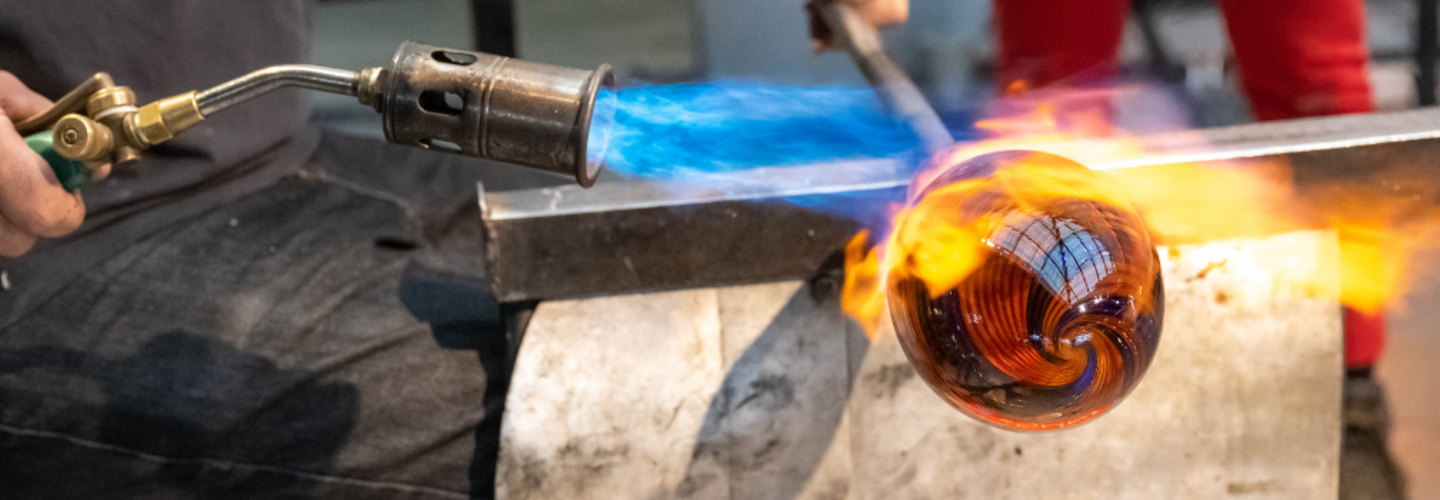 Man holds blow torch to glass art piece that is fixed to the end of a glass blowing stick.