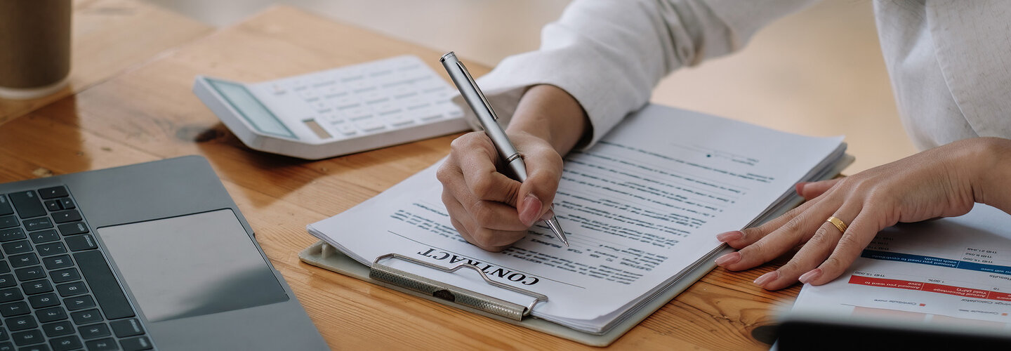 Hand signing contract with pen near laptop.