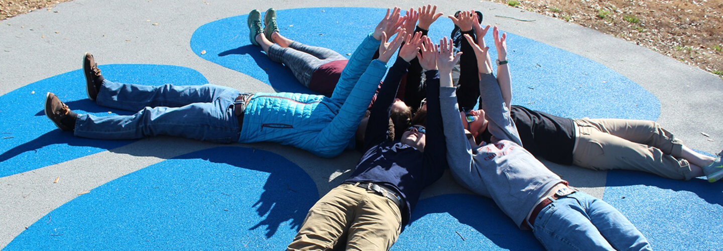 Students lying on the concrete in a circle with their shoulders touching and arms up in the air with palms facing out. 