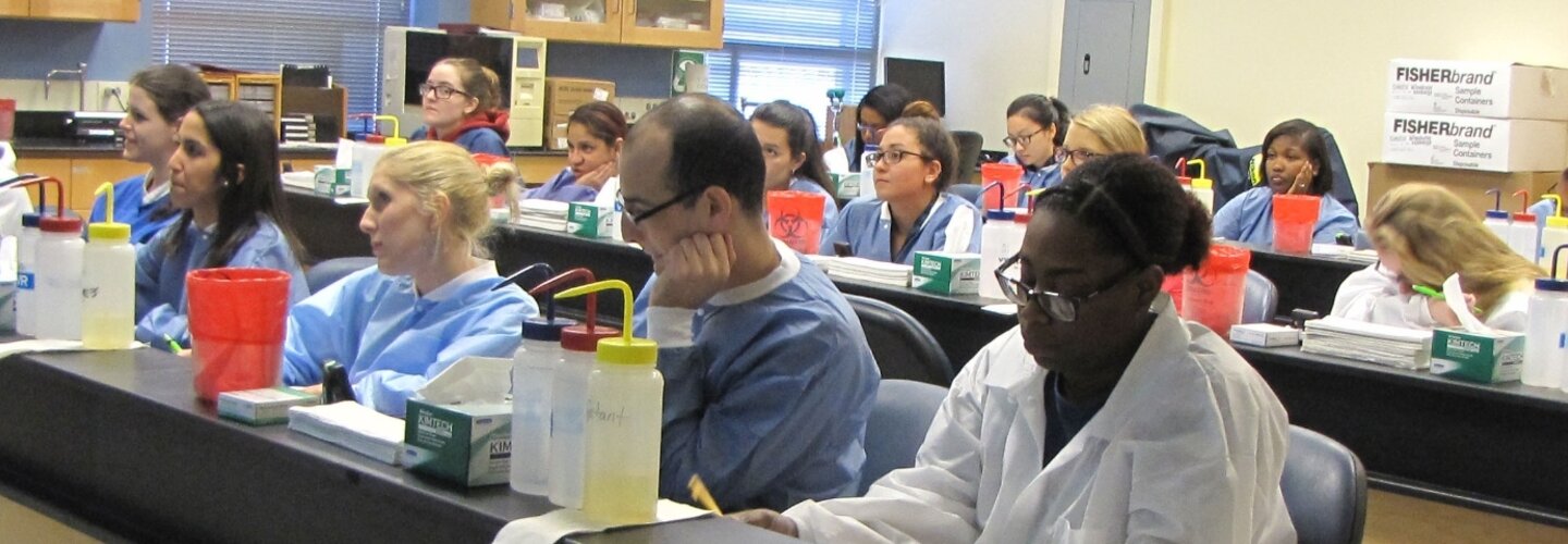 science students sitting in scrubs with different lab materials next to each of them.