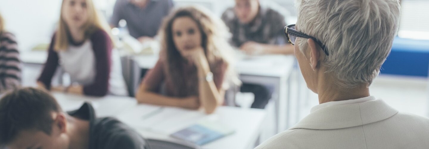 Back view of grey-haired woman teacher stading at classroom and talking to high school students.