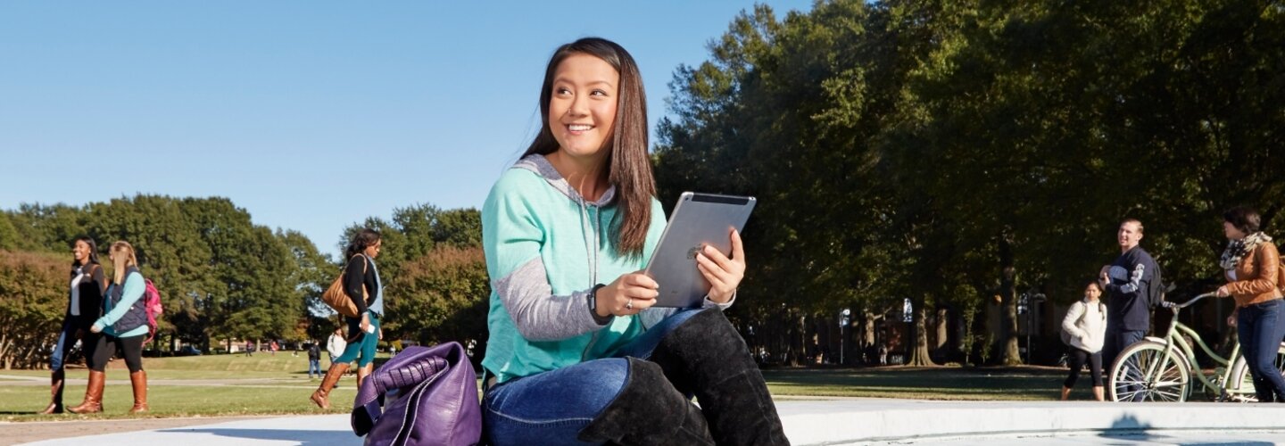 A smiling female student sit at the fountain holding a tablet while students walk past.