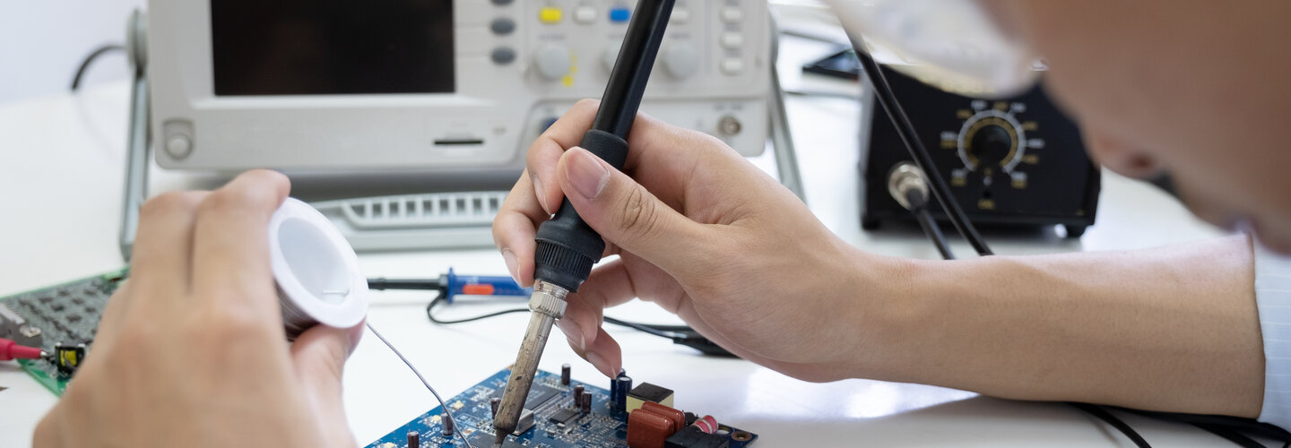 Electrical engineering banner image of student using a soldering tool on an electric panel. 
