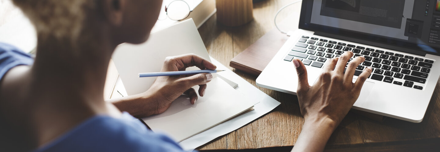 women typing on computer with a pen in her other hand on a pad of paper.