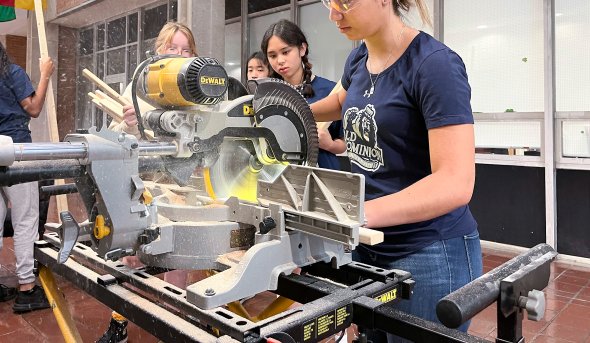 A young woman in T-shirt, jeans and goggles cuts wood with an electric saw.