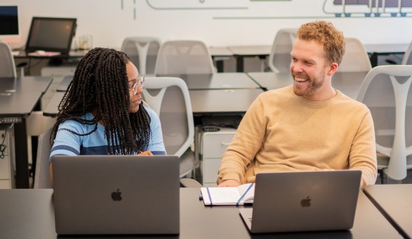 A female student laughs with a male supervisor in an office space while they look at laptops.