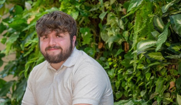  Adyn, a young White man with brown hair and beard wearing a beige collared shirt, sits against a background of greenery. 