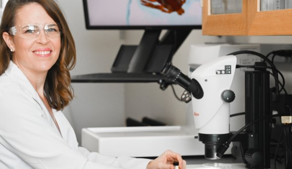 A smiling woman in a lab coat sits at a laboratory desk with a large picture of a tick behind her.