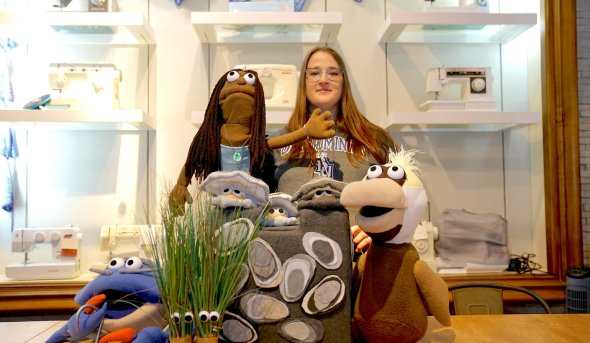 Sydney Walker poses with puppet characters from “The Amazing Neighborhood Mollusk Adventure” including Mr. Blue the crab, Reign the mermaid, a trio of oysters called the Reef Rascals and Esperanza, a brown pelican. Two clumps of marsh grass with googly eyes, named Tina and Flora, stand in the foreground.