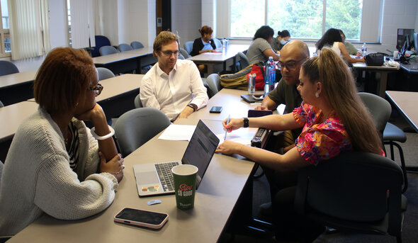 A group of students collaborate around a table in a classroom, with laptops, papers and a coffee cup on the surface