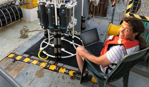 A woman sits on a ship next to a piece of research equipment. 