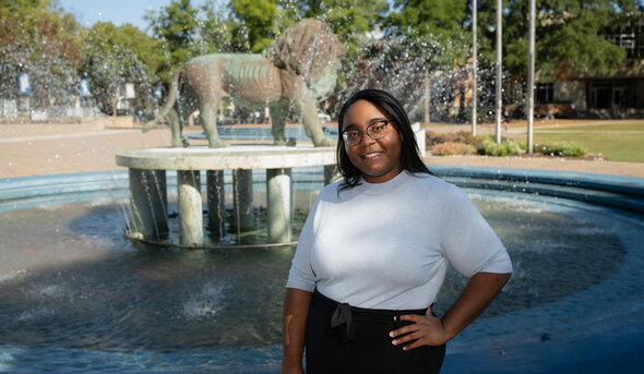 Destini Harrell, smiling at the camera, stands in front of the ODU lion fountain silhouetted against a blue sky.