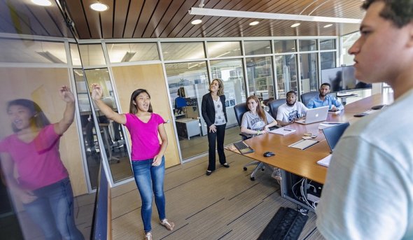 Students in conference room sit at table while looking at whiteboard.