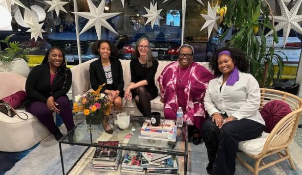 Five women sitting on chairs around a coffee table smile at the camera