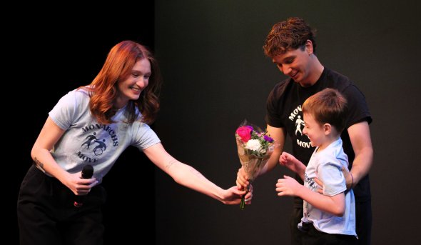 Baillie Ollila, a young woman with red hair wearing a Monarchs Move t-shirt, hands a young dance participant flowers, alongside another volunteer.