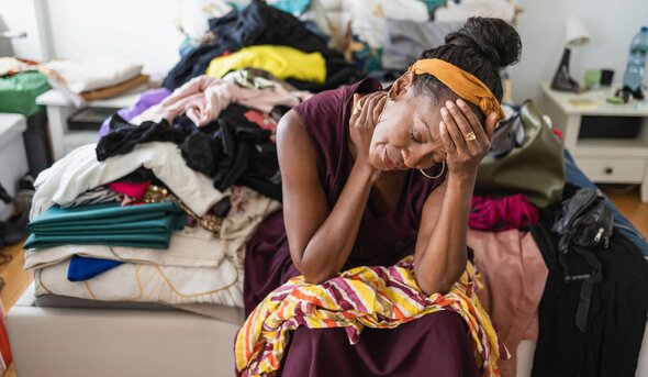 Overwhelmed woman  sitting on a messy bed covered with clothes with her head leaning on her hand.