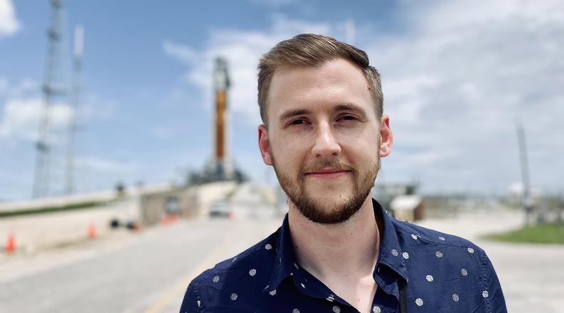 a headshot style photo of Ian Haskin. He is wearing a blue button-up shirt and standing in a NASA airfield