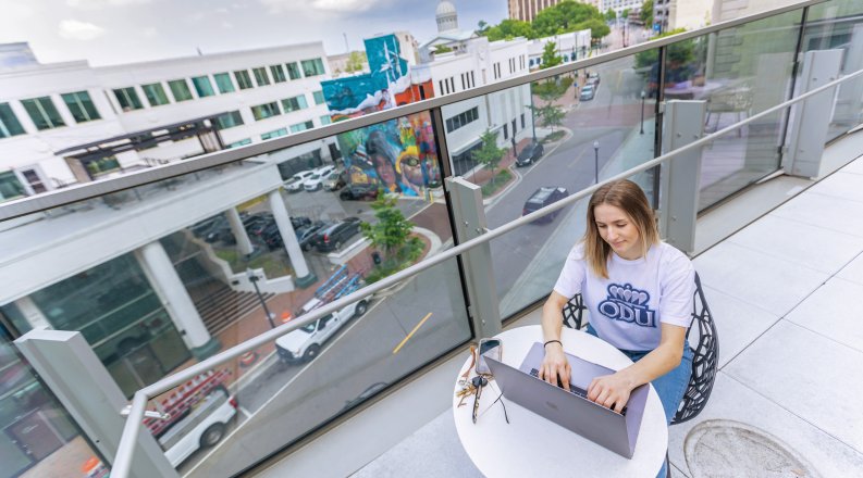 a female student with blond hair and a white odu t-shirt is sitting on a rooftop in a downtown area studying