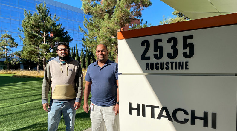 two men pose in front of the hitachi sign