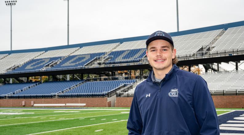 a male students wearing a baseball hat stands in front of the football field