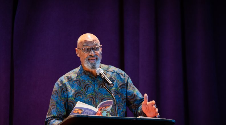 A man reads from a book during a presentation at a lectern.