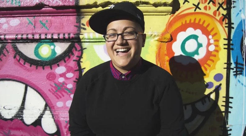 Artist Gabby Rivera stands in front of a colorful mural. 