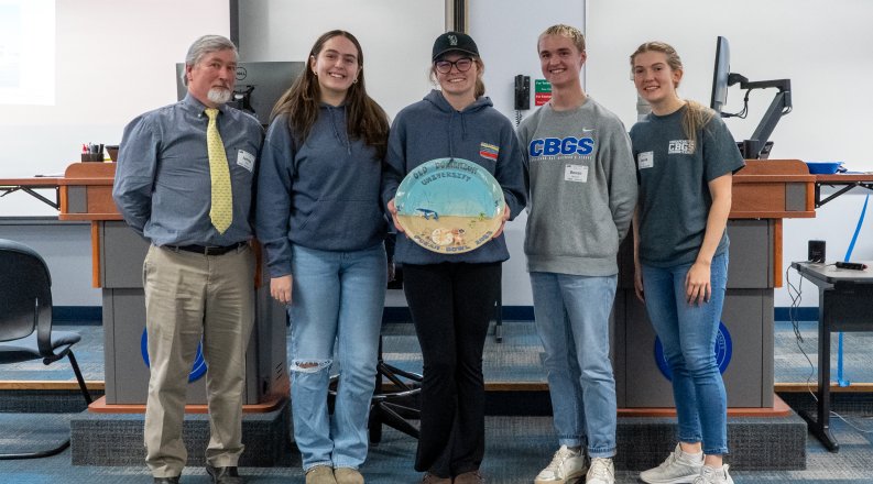 A group of students and their teacher stand with an oval-shaped bowl recognizing their first-place finish in the first-ever Ocean Bowl competition.  