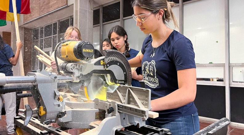 A young woman in T-shirt, jeans and goggles cuts wood with an electric saw.
