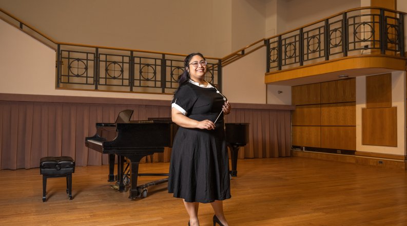 A woman stands in front of a piano, smiling and holding a conductor's baton.