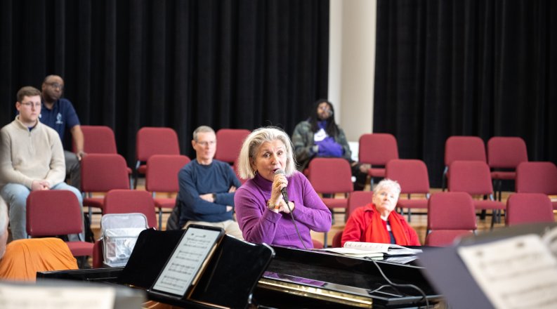 A woman sings into a microphone with audience members in the background. 