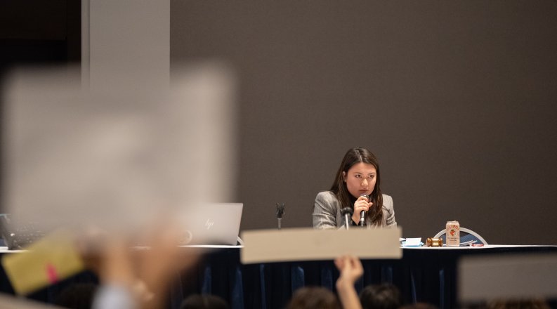 A woman sits at a table and speaks into a microphone.