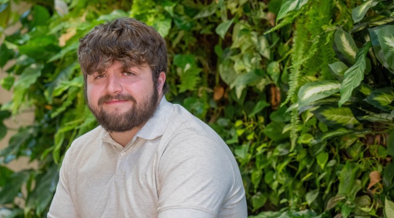  Adyn, a young White man with brown hair and beard wearing a beige collared shirt, sits against a background of greenery. 