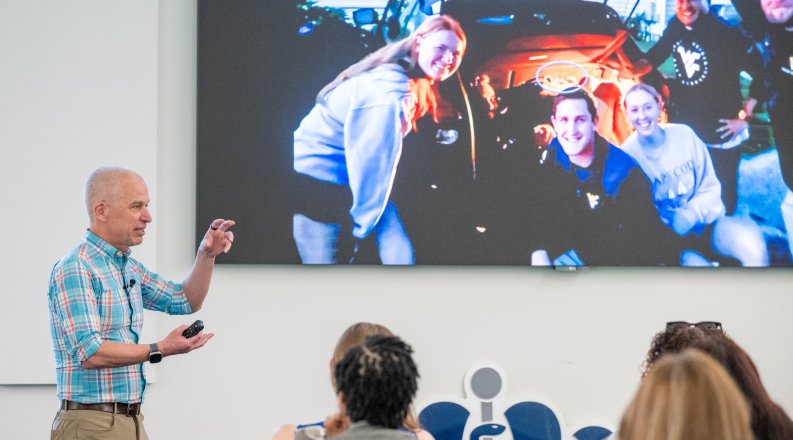Douglas Casa stands in a lecture hall gesturing with his hands towards a screen