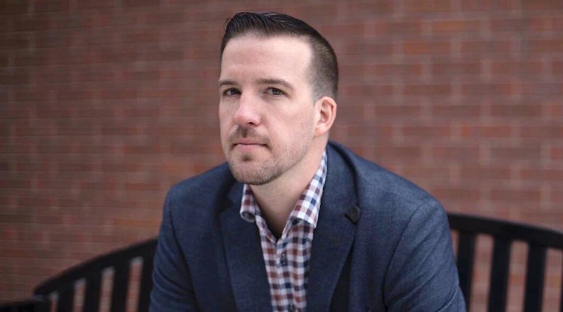 Larson dressed in a blue suit standing in front of brick wall. 