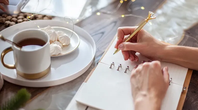 A woman writes in a blank notebook with tea and cakes on a t