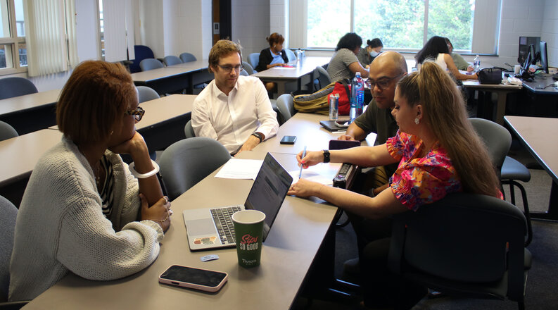 A group of students collaborate around a table in a classroom, with laptops, papers and a coffee cup on the surface