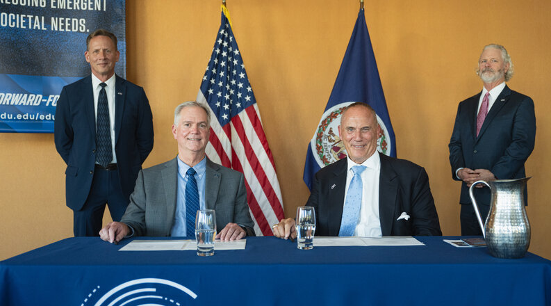 two men seated at table two stand stand behind flanking flags