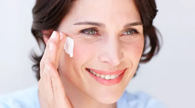 A mature woman with wavy brown hair smiles as she applies mo
