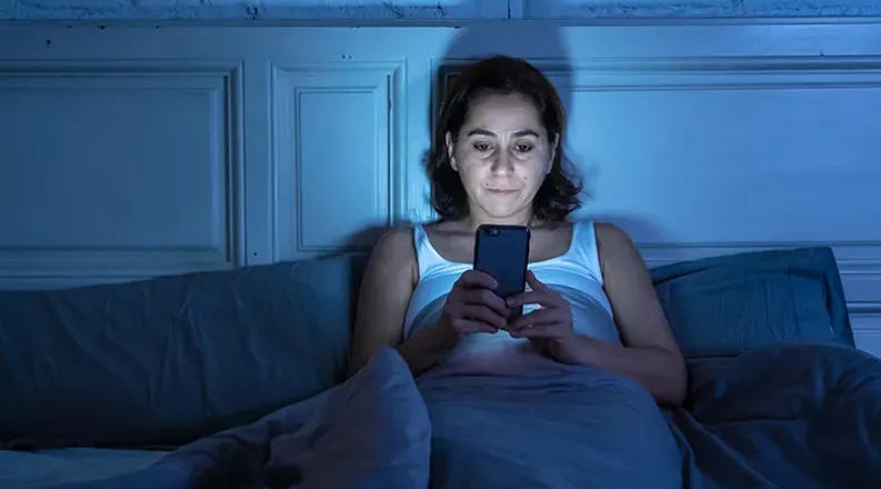A woman sits up in bed in a dark room, looking at her cell p